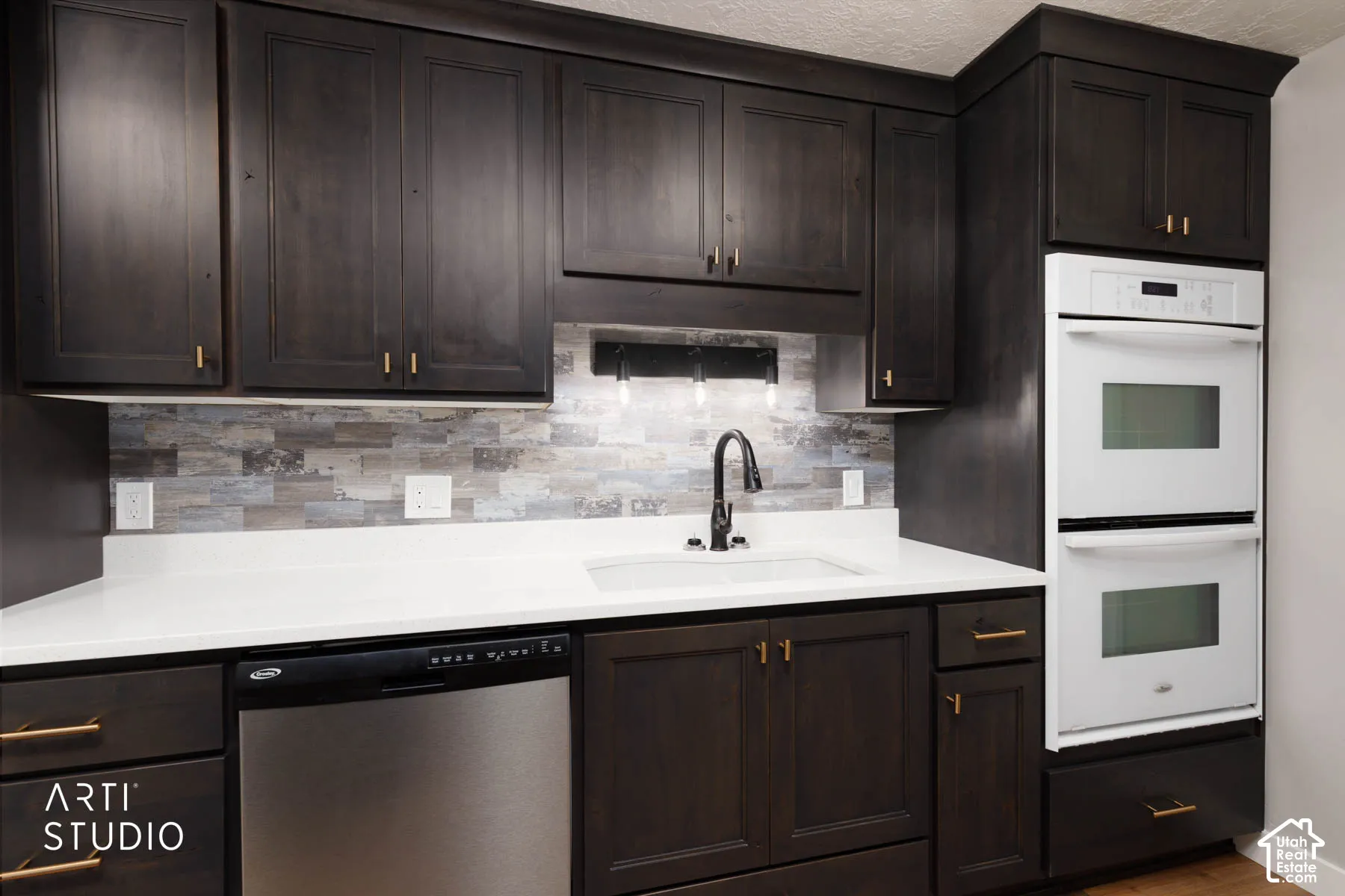 Dark-stained cabinetry with gold-toned hardware, white solid surface countertops, mosaic tile backsplash, integrated stainless steel dishwasher, and built-in double ovens