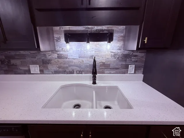 White double-basin sink set into a speckled white countertop, accented by a dark faucet and a stone-look tile backsplash