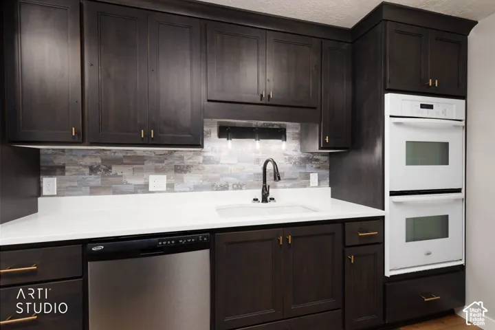 Dark-stained cabinetry with gold-toned hardware, white solid surface countertops, mosaic tile backsplash, integrated stainless steel dishwasher, and built-in double ovens