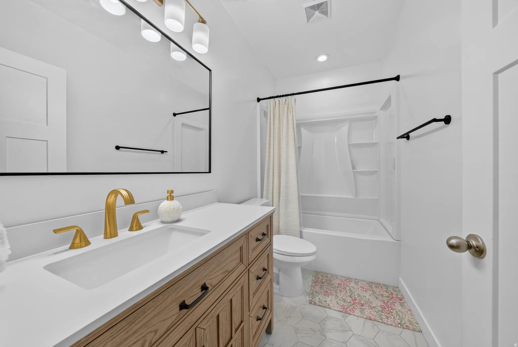 Wood-finish vanity with a white countertop and rectangular undermount sink, gold-tone faucet, large rectangular mirror, and hexagonal floor tiling