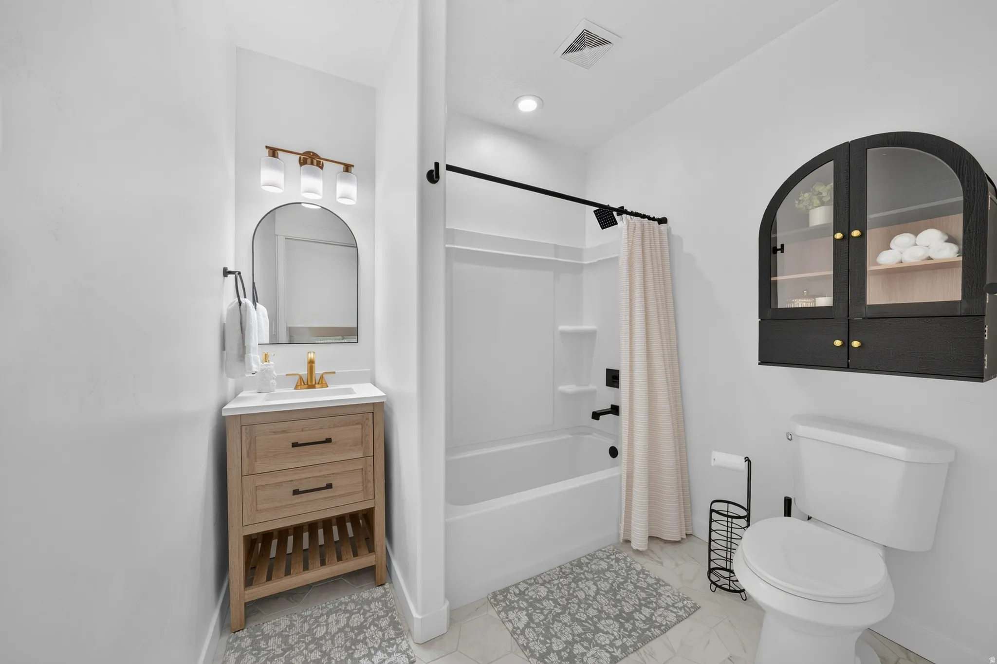 Wood-finish vanity with a white countertop and gold-tone faucet, arched mirror, and a built-in shower and tub combination