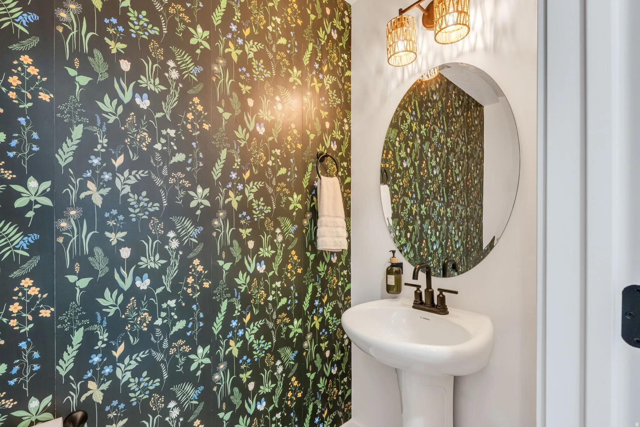 Botanical pattern wall covering accent wall, pedestal sink with dark bronze faucet, round wall-mounted mirror, and decorative wall sconce lighting