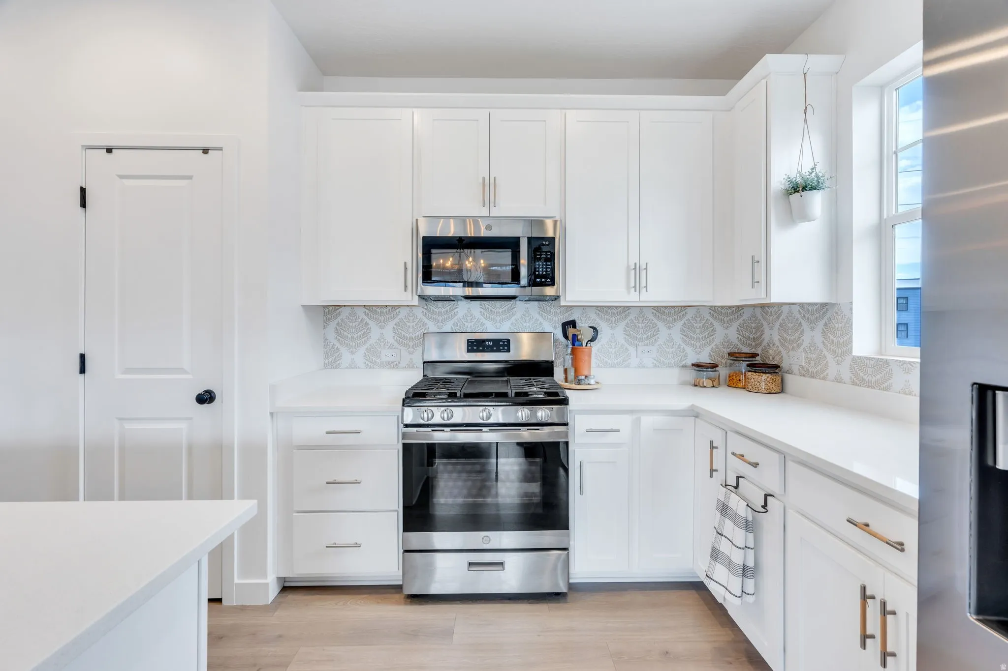 White shaker cabinetry, light-toned wood-finish flooring, stainless steel range with overhead microwave, solid surface countertops, and patterned tile backsplash