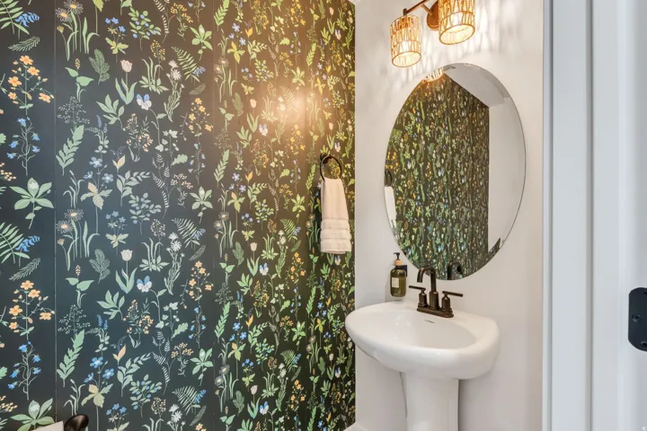 Botanical pattern wall covering accent wall, pedestal sink with dark bronze faucet, round wall-mounted mirror, and decorative wall sconce lighting