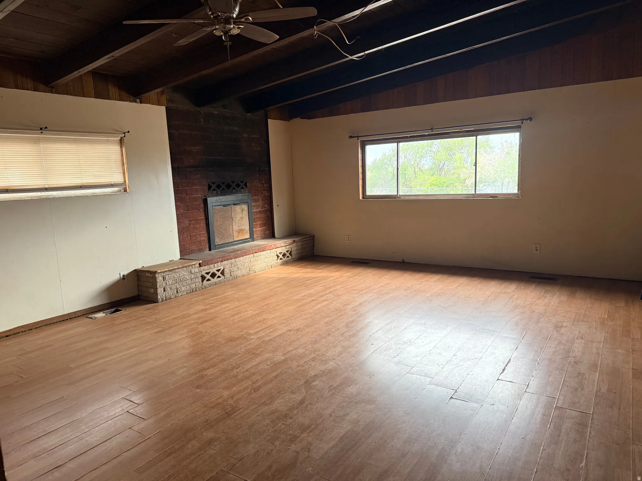 Wood-finish flooring, exposed wood beams, a brick fireplace with a hearth, and a ceiling fan