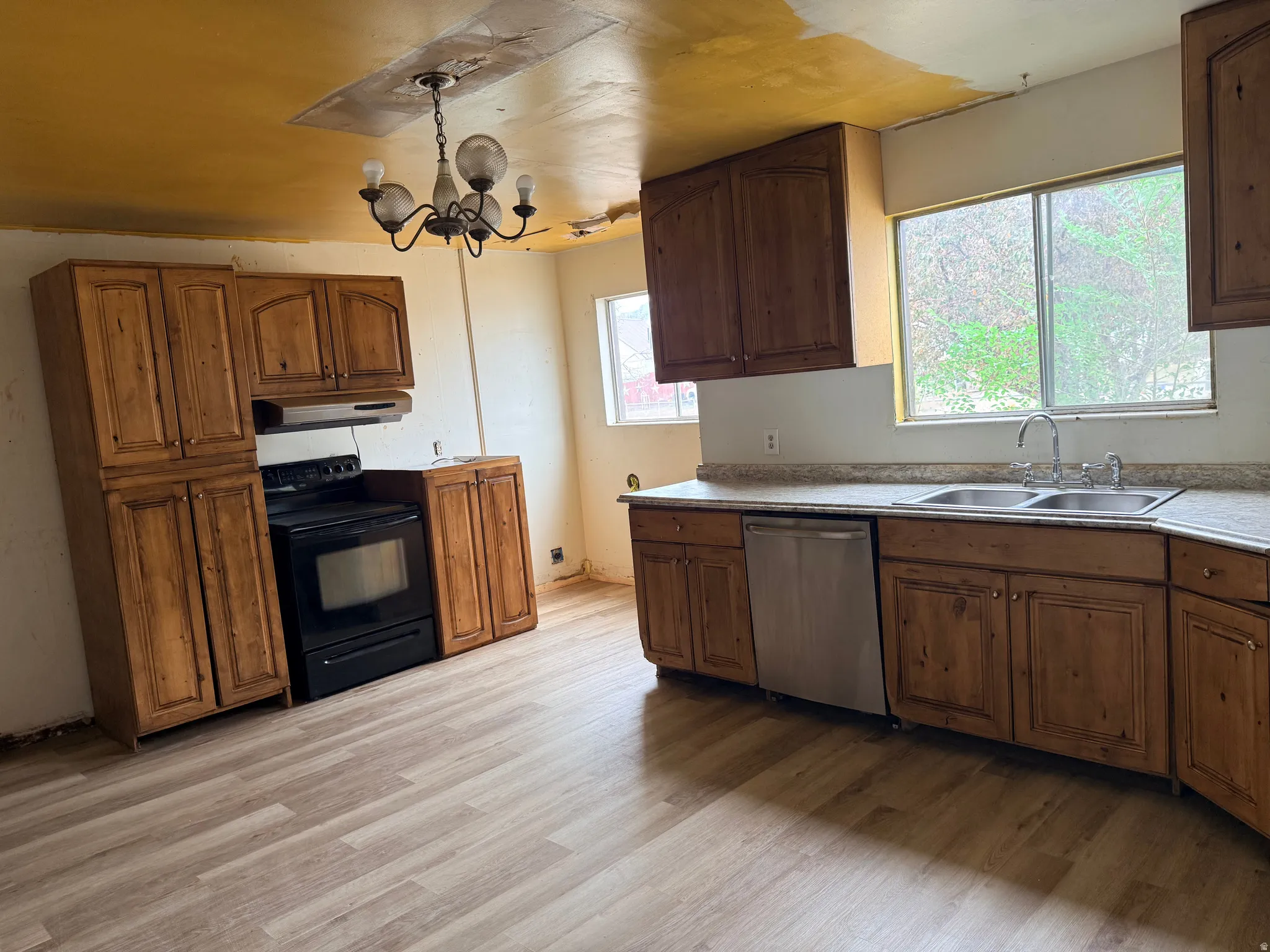 Wood-finish flooring, ample wood cabinetry, double basin stainless steel sink, stainless steel dishwasher, and a black range with an overhead vent