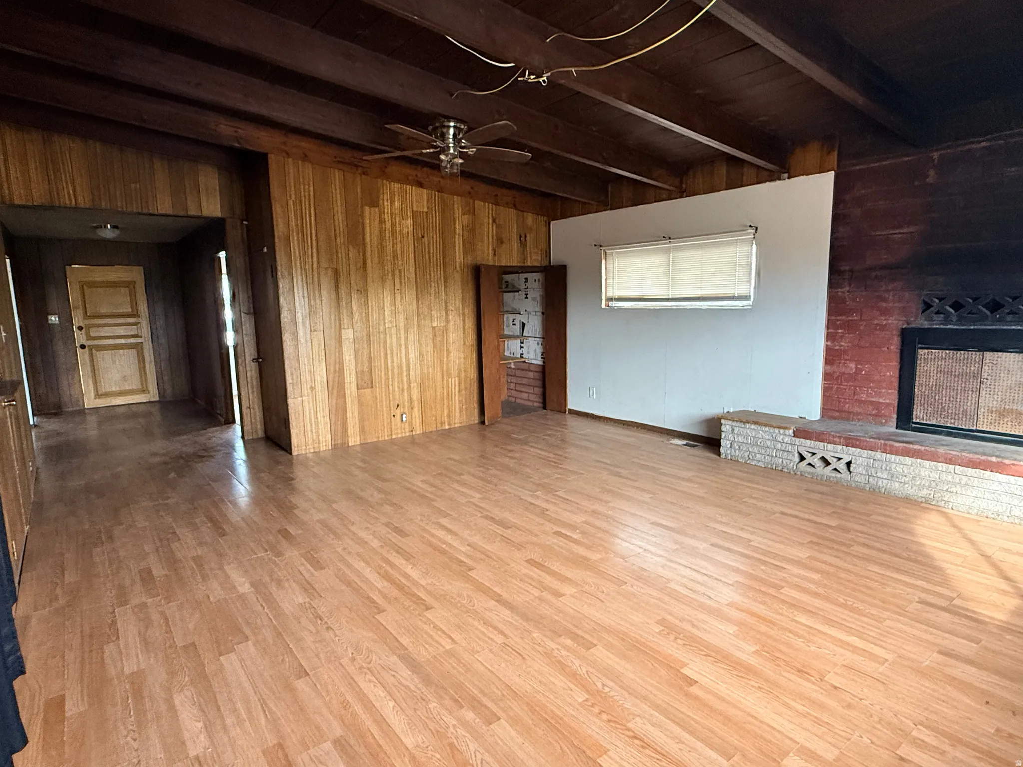 Wood-finish flooring throughout, exposed wood beam ceiling, wood paneled walls, brick fireplace with hearth, and a ceiling fan