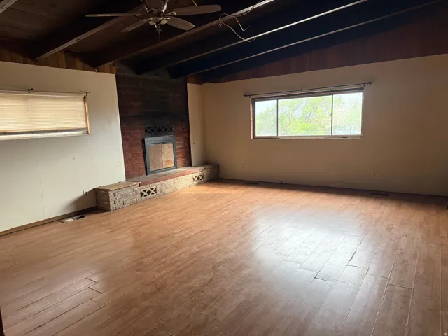 Wood-finish flooring, exposed wood beams, a brick fireplace with a hearth, and a ceiling fan