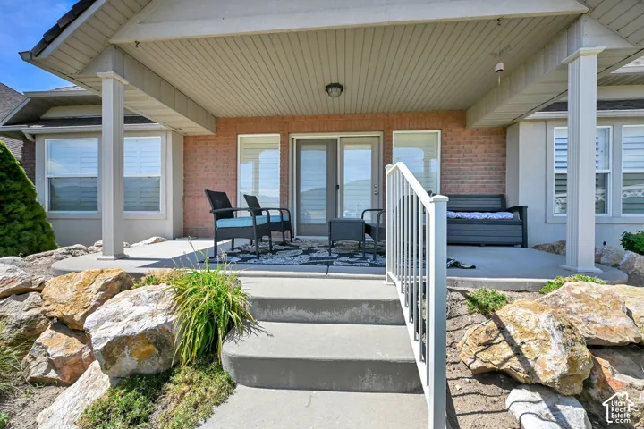 Covered concrete patio with overhead lighting, brick exterior wall, sliding glass door, and a white railing leading to the lower level