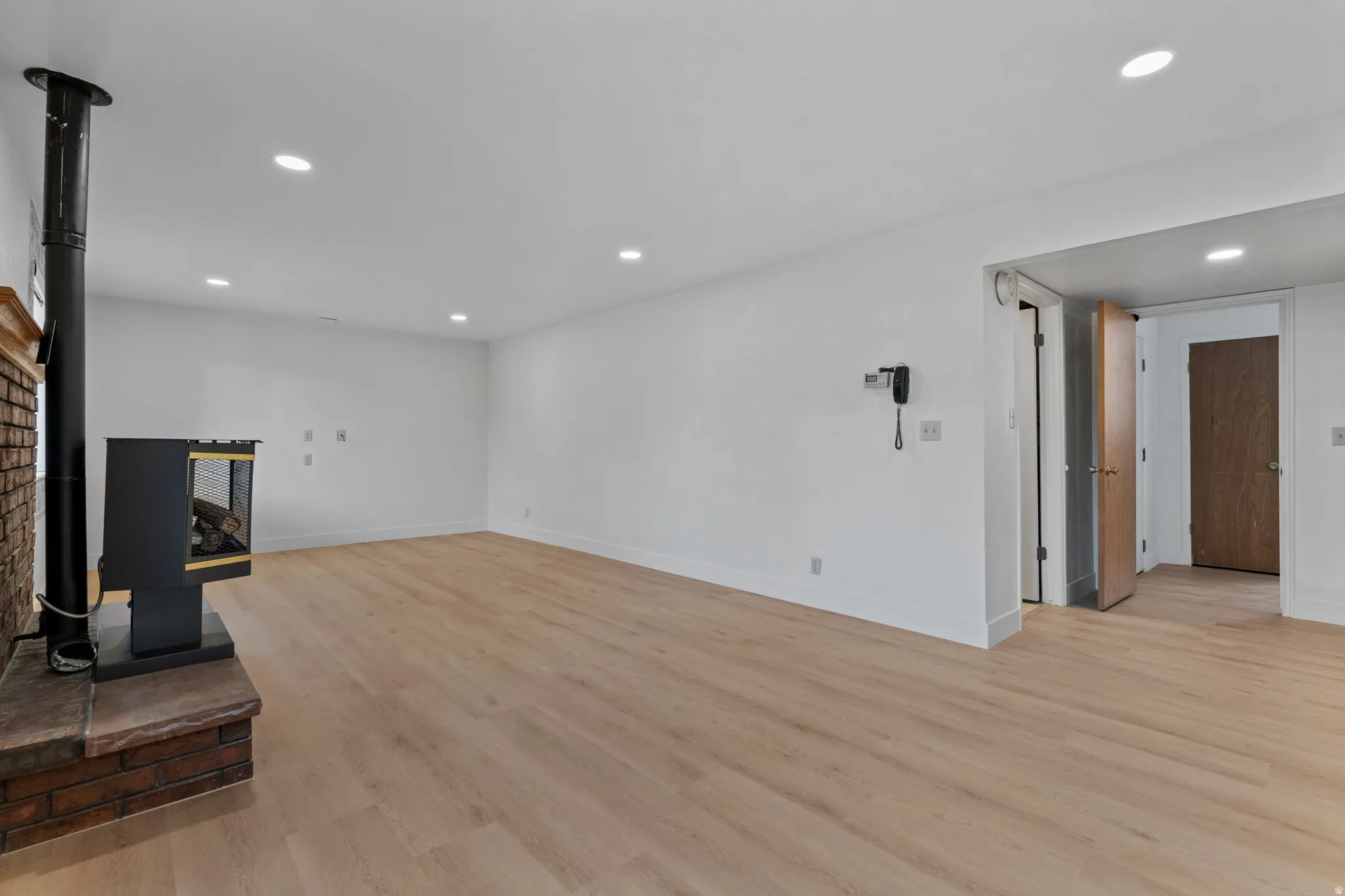 Expansive living area featuring light wood-finish flooring, recessed lighting, a freestanding fireplace with a brick hearth, and crisp white walls