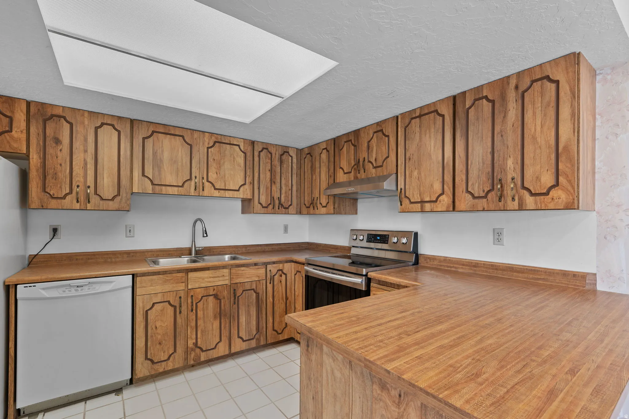 Wood-finish cabinetry, stainless steel range with overhead ventilation, double basin sink with gooseneck faucet, white dishwasher, and tiled flooring