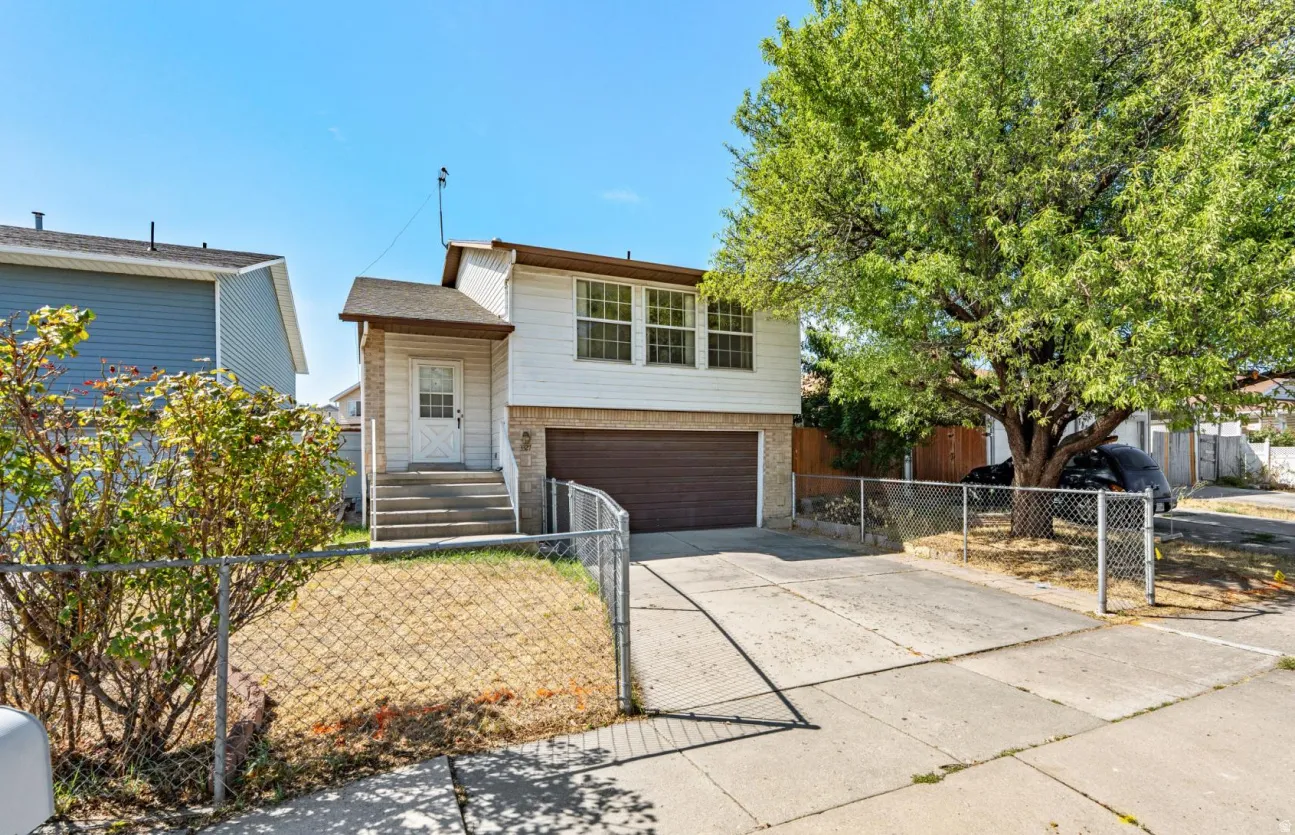 White siding exterior with a brown garage door, front entry stairs, a chain link fence, and a mature tree