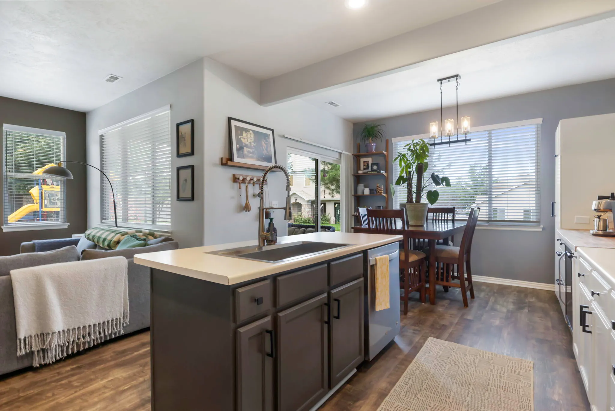 Open-concept living area with wood-finish flooring, a kitchen island with sink, and a dining area featuring a linear chandelier