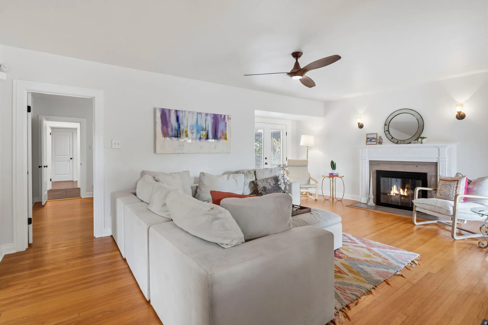 Hardwood flooring, white painted walls, fireplace with white mantel, ceiling fan, and recessed alcove with French doors