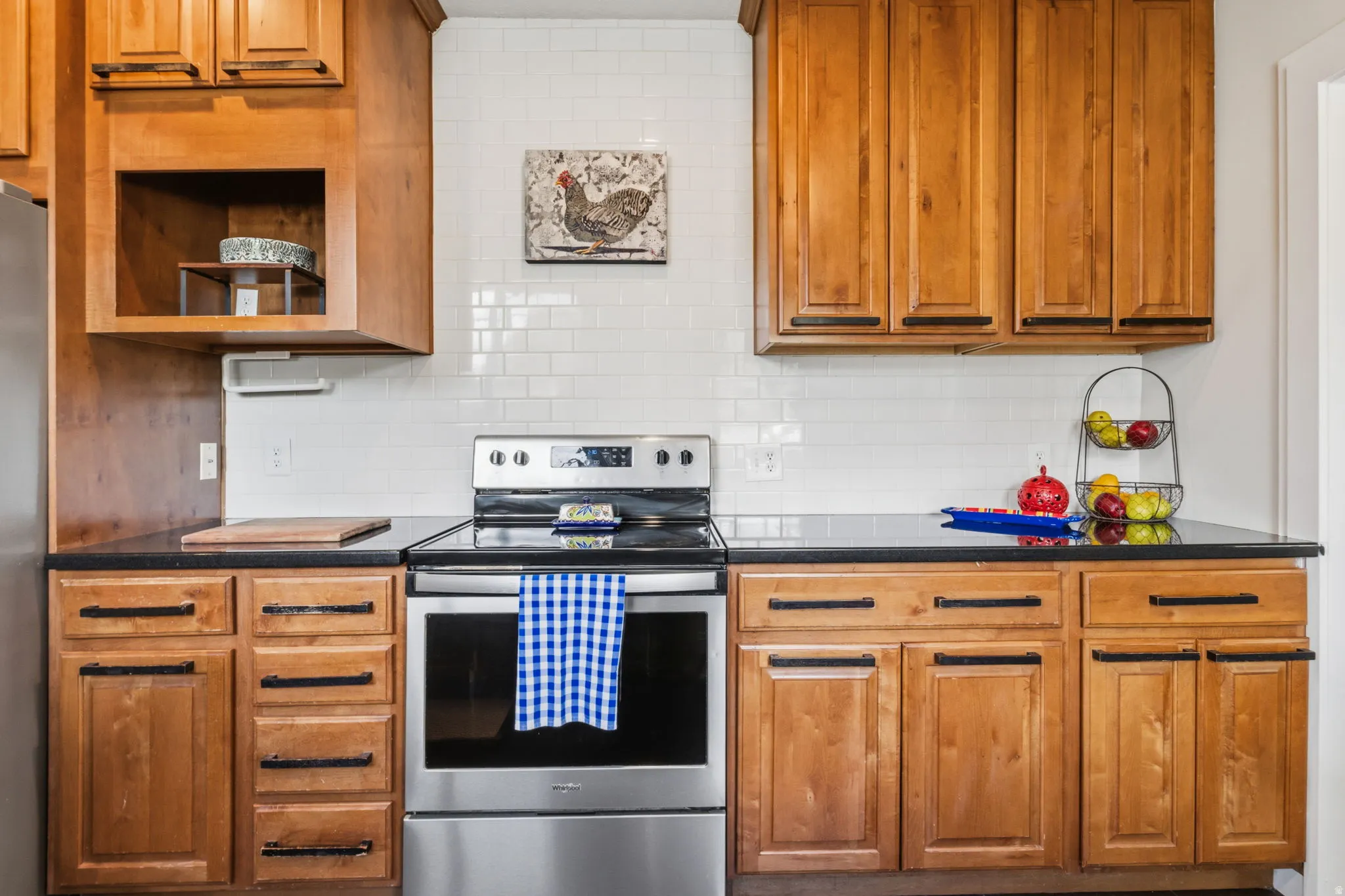 Wood-finish cabinetry with black hardware, black countertops, stainless steel range, and white subway tile backsplash