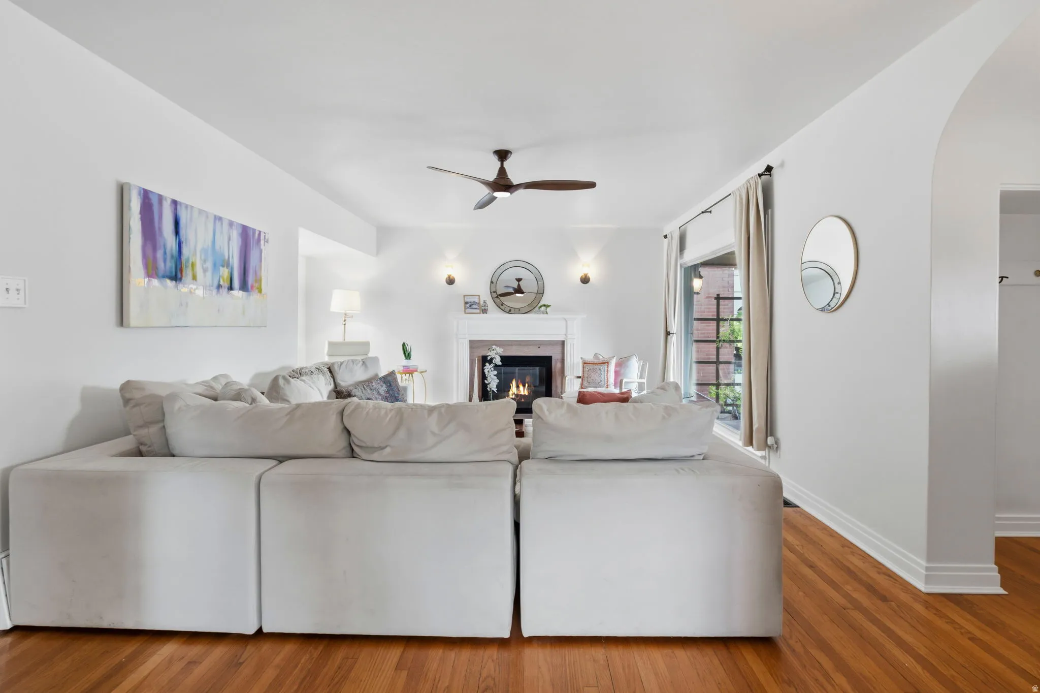 Hardwood flooring, arched doorway, fireplace with mantel, ceiling fan, and large window with exterior access