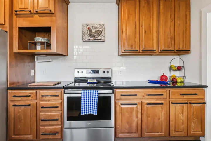 Wood-finish cabinetry with black hardware, black countertops, stainless steel range, and white subway tile backsplash