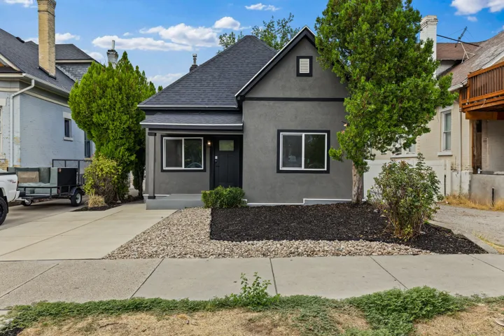Stucco exterior with contrasting trim, dark shingle roof, paved driveway, and mature landscaping