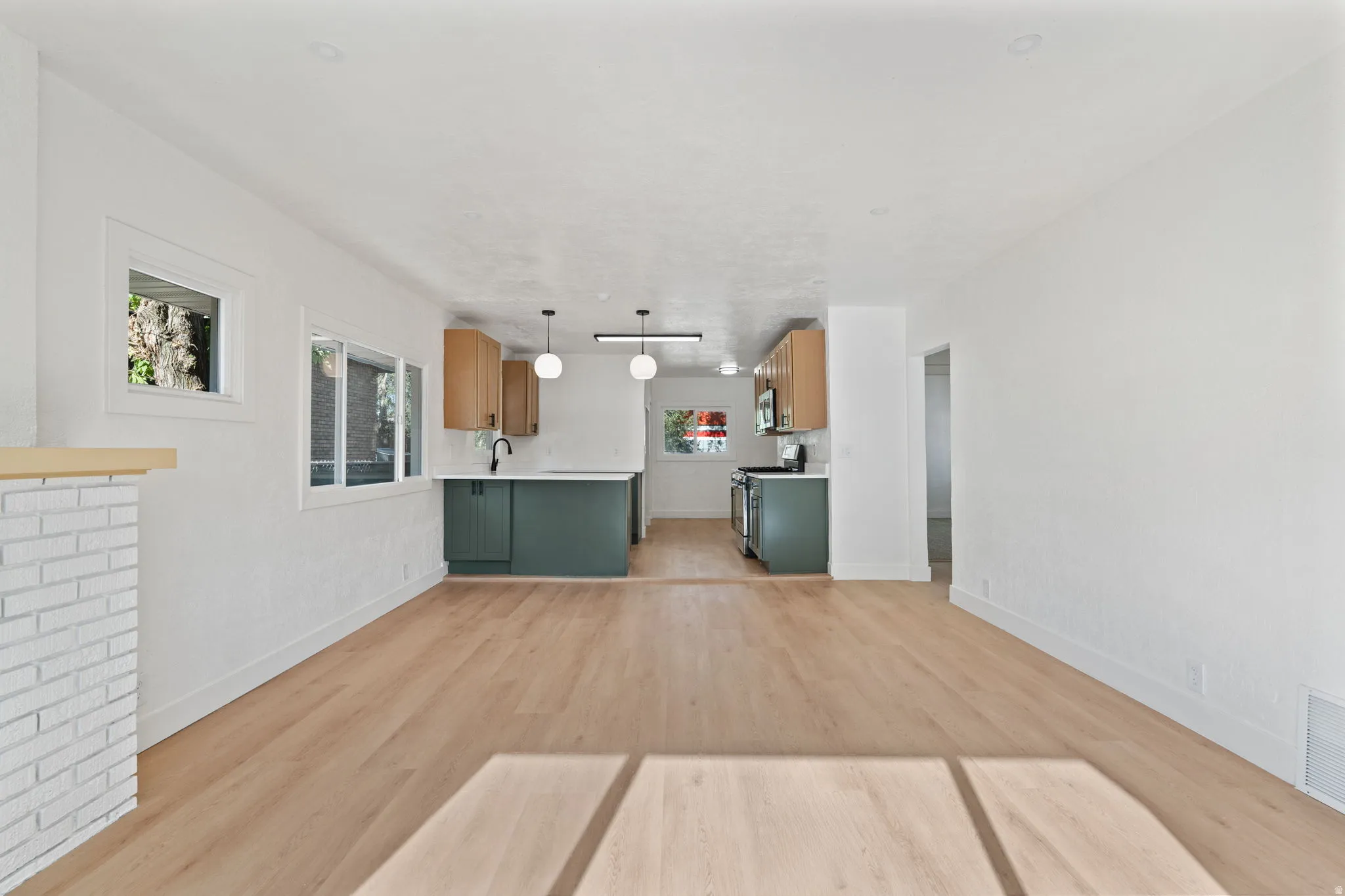 Wood-finish flooring throughout, white painted walls, white brick fireplace with a wood mantel, and a kitchen featuring dark green lower cabinetry and wood-finish upper cabinetry