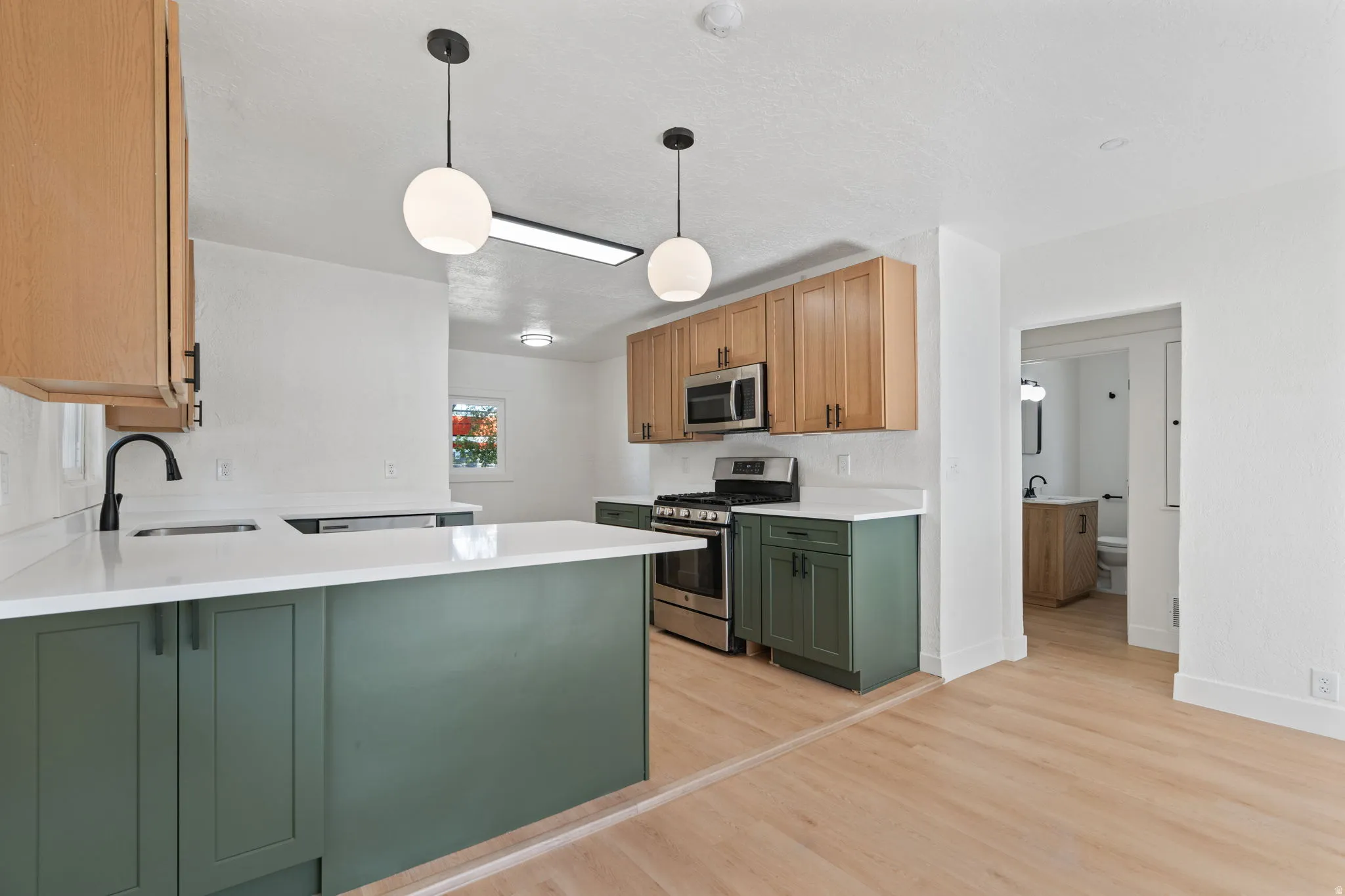 Kitchen featuring two-tone cabinetry with wood-finish upper cabinets and green lower cabinets, light-toned countertops, stainless steel appliances, and a built-in sink with a matte black faucet