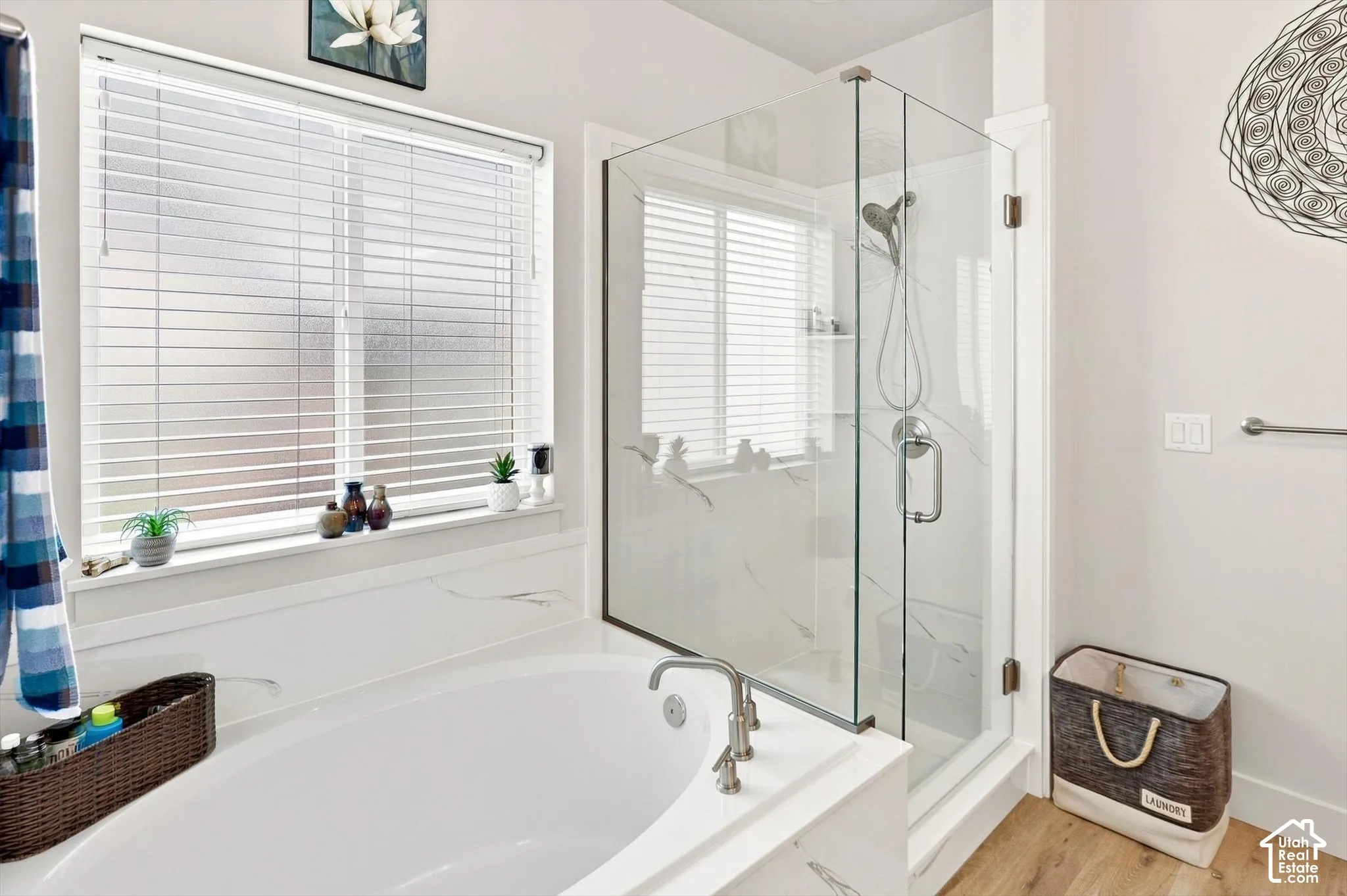 Freestanding soaking tub with deck-mounted faucet, glass-enclosed shower, large window with privacy blinds, and wood-finish flooring