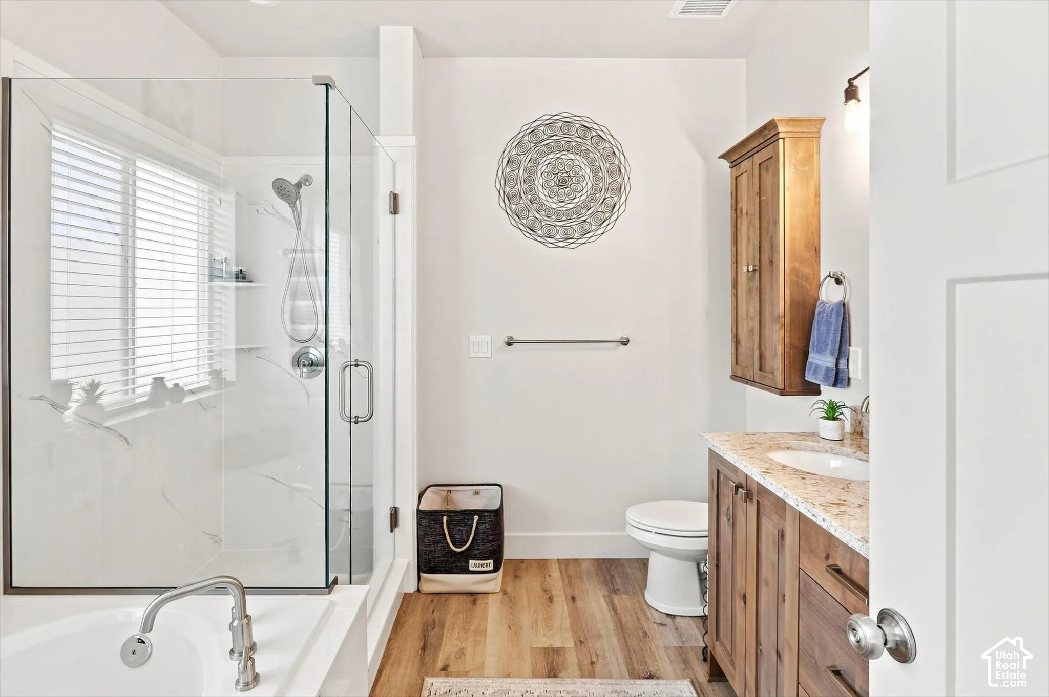 Glass-enclosed shower with white tiled walls, separate bathtub with polished chrome fixtures, wood-finish flooring, wood vanity with granite-style countertop, and built-in wood wall cabinet