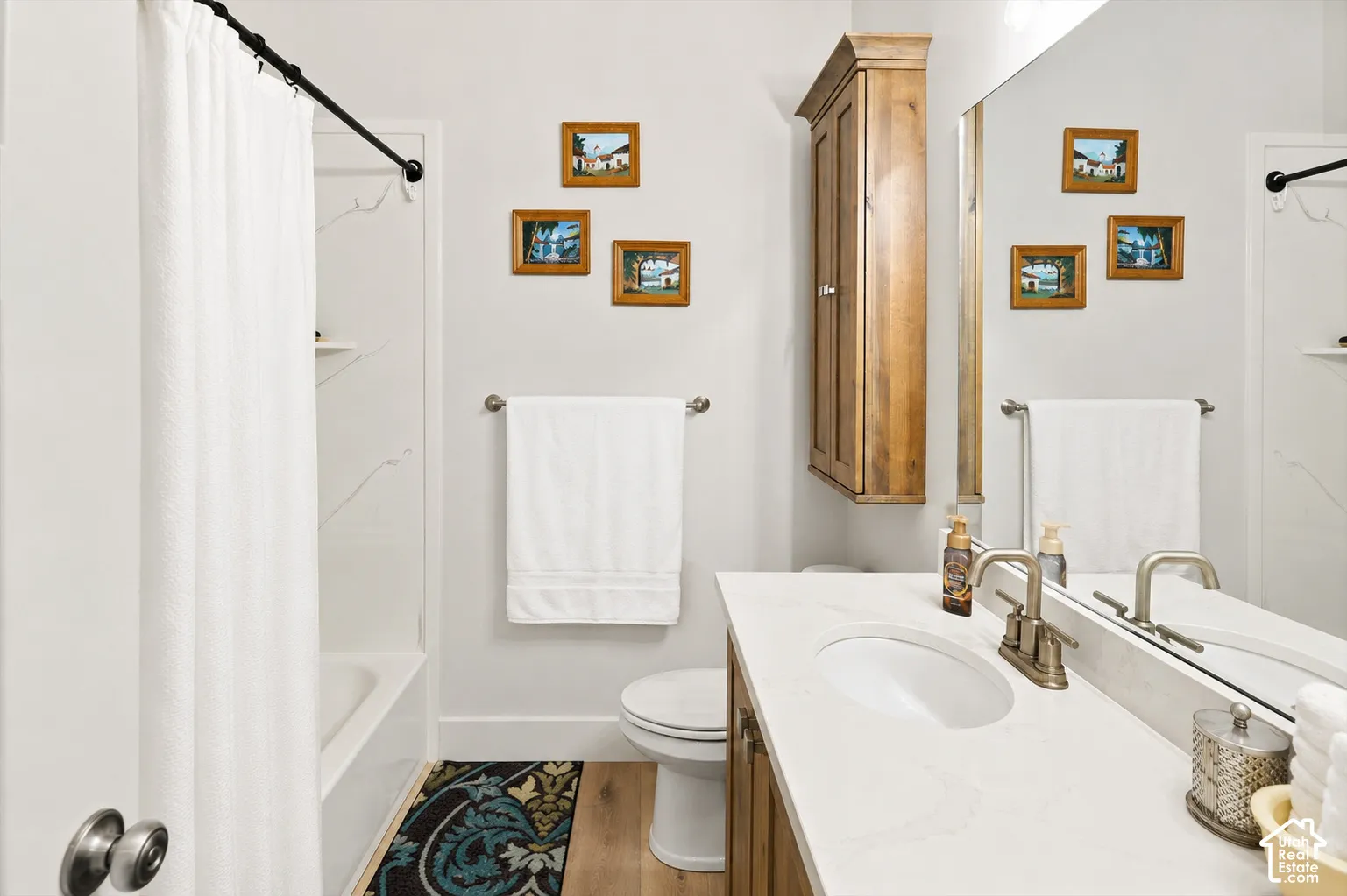 Integrated shower-over-tub with white surround, single vanity with countertop, wood-finish base cabinetry, wall-mounted wood-finish cabinet, and wood-finish flooring
