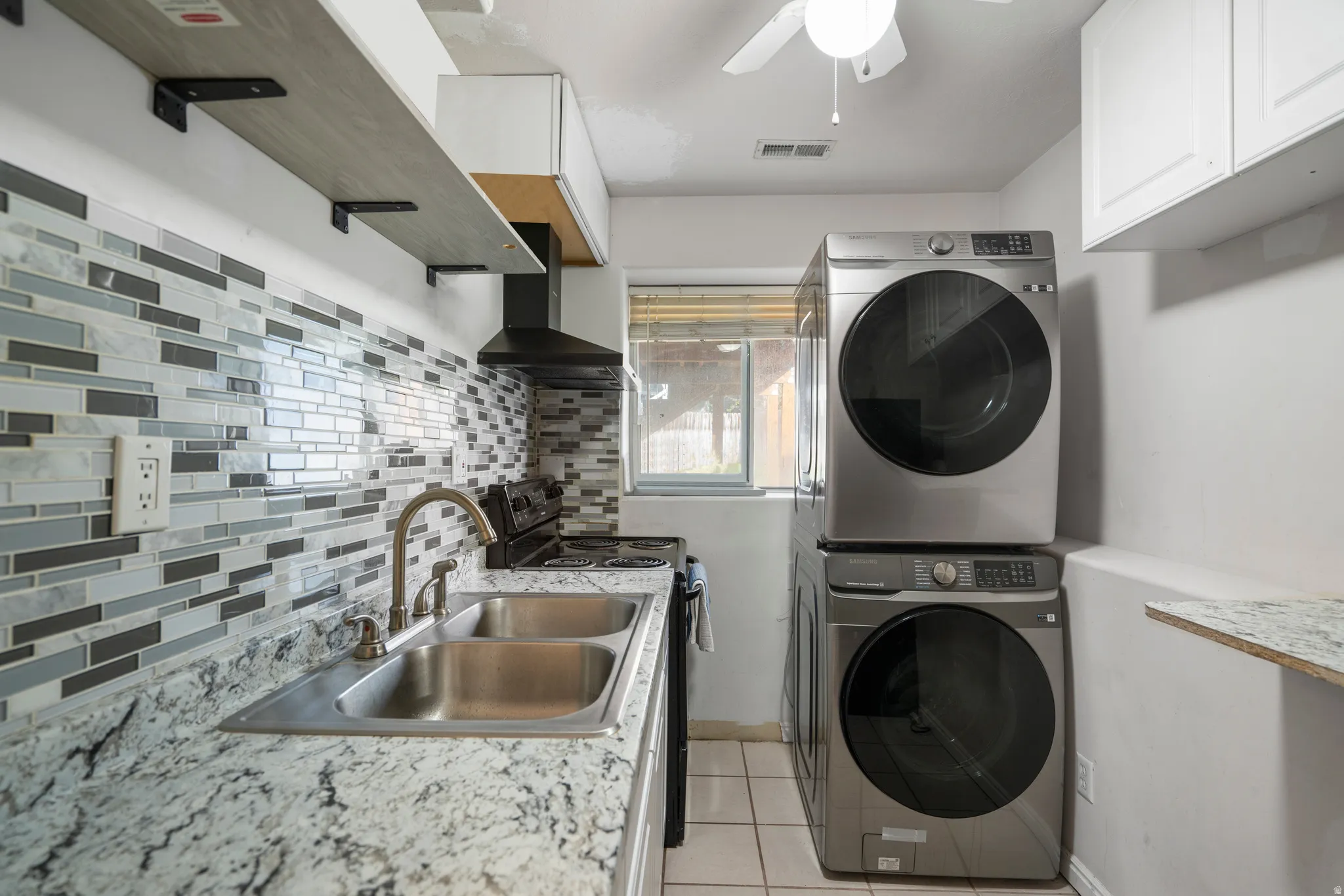 Full-height backsplash with a mosaic tile pattern, double basin sink with a brushed nickel faucet, light-toned countertop, open shelving with black brackets, and a stacked washer and dryer