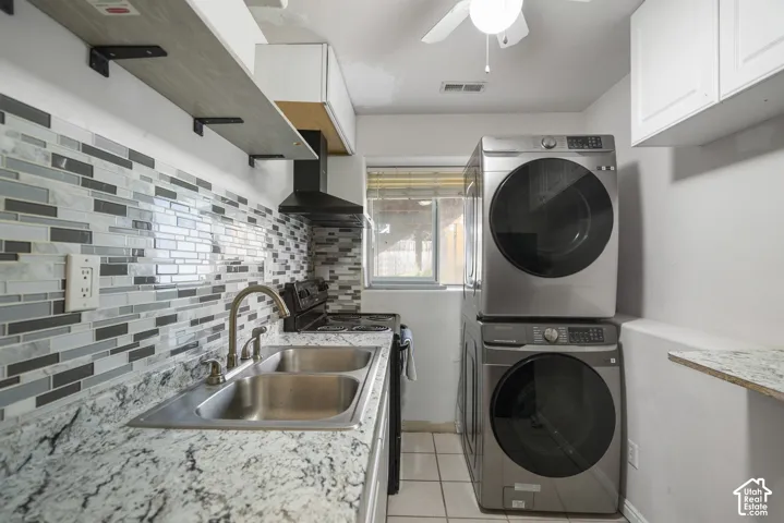 Full-height backsplash with a mosaic tile pattern, double basin sink with a brushed nickel faucet, light-toned countertop, open shelving with black brackets, and a stacked washer and dryer