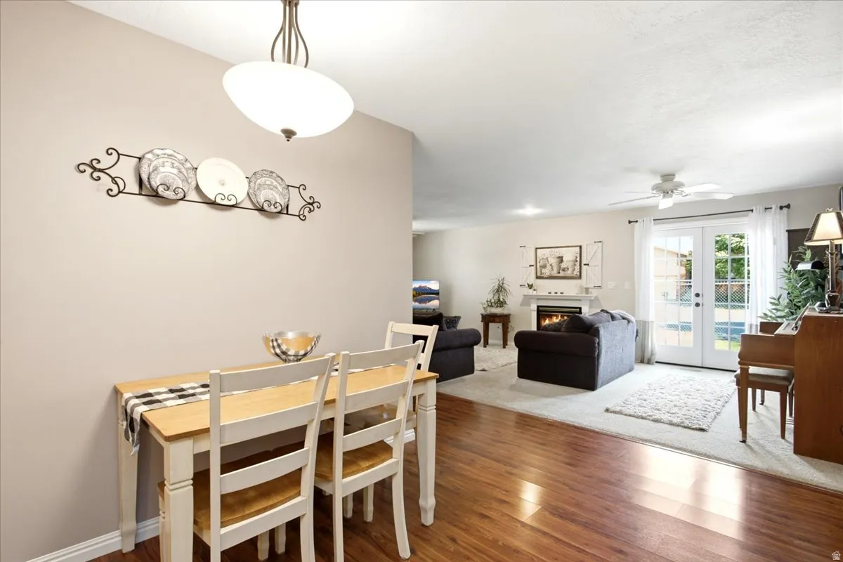 Open-concept living area featuring warm wood-finish flooring, a dining table with seating for four, and a spacious adjoining living room with recessed ceiling lighting and glass doors providing natural illumination
