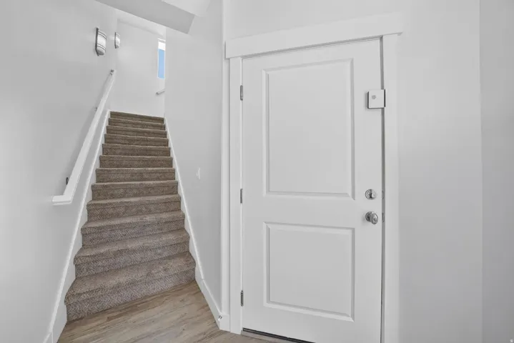 Entryway with wood-finish flooring, two-panel interior door, and carpeted staircase featuring a white handrail and wall-mounted sconce lighting