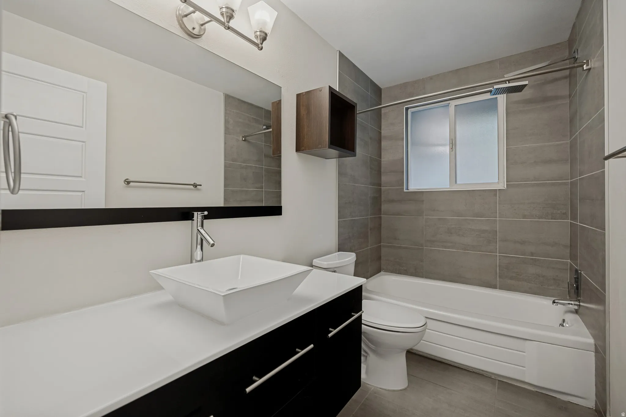 Contemporary bathroom featuring a floating vanity with a vessel sink, a full-width mirror, tile flooring, a bathtub with a shower system, and a window with privacy glass