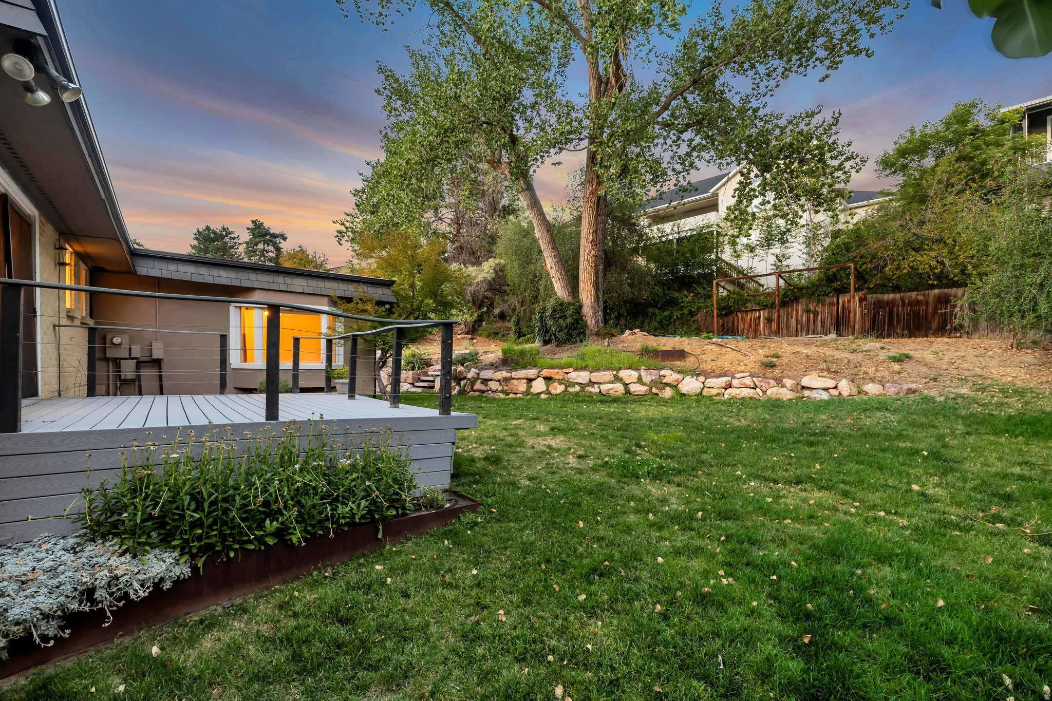Expansive backyard featuring a large deck with cable railings, manicured lawn, and natural stone retaining walls