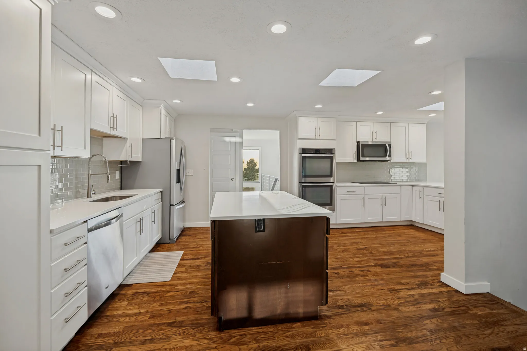 Spacious kitchen featuring white cabinetry, stainless steel appliances including double wall ovens and a gas cooktop, a large center island with white quartz countertop, subway tile backsplash, recessed lighting throughout, and warm wood-finish flooring