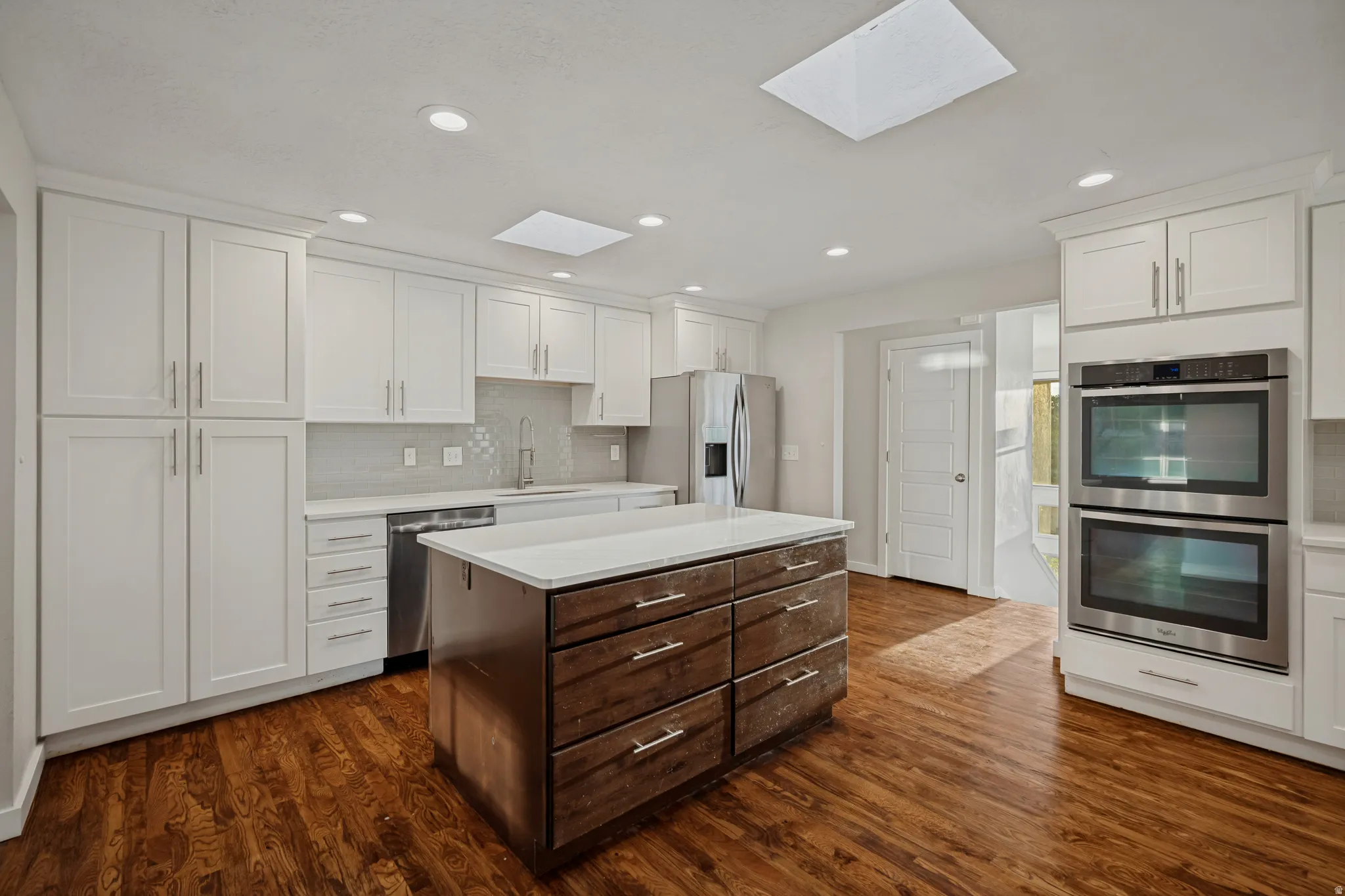 Kitchen featuring white shaker cabinetry, stainless steel appliances, wood-finish flooring, a center island with countertop, and skylights