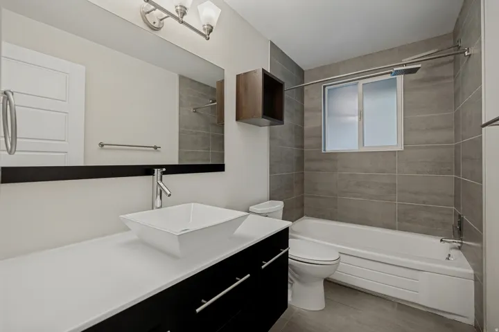 Contemporary bathroom featuring a floating vanity with a vessel sink, a full-width mirror, tile flooring, a bathtub with a shower system, and a window with privacy glass