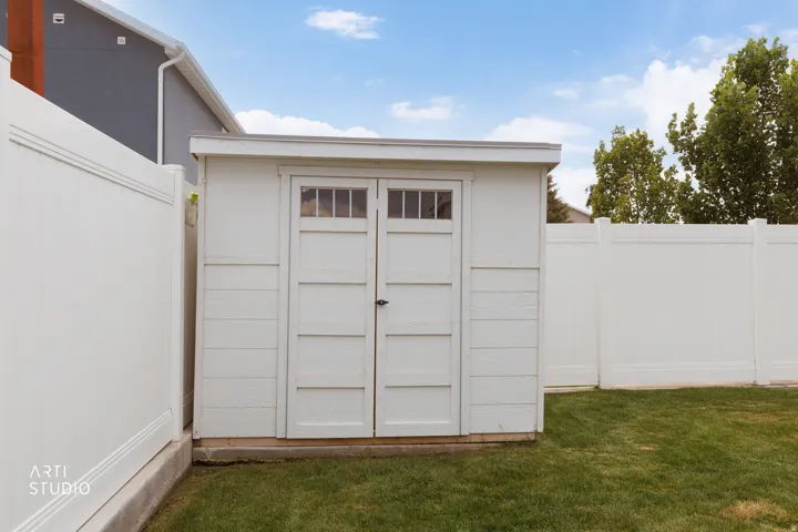White storage shed with transom windows, surrounded by a white privacy fence and a manicured lawn