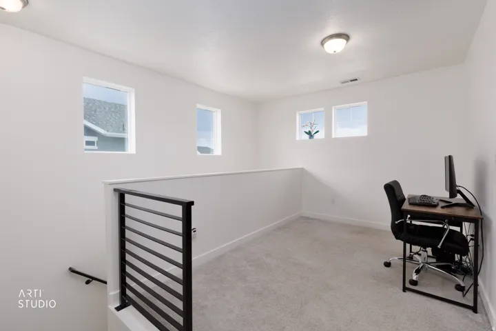 Open loft area featuring light gray carpeting, crisp white walls, multiple windows, and a contemporary black metal stair railing