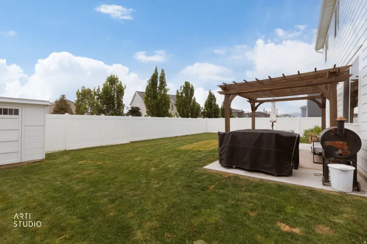 Expansive green lawn, white vinyl fencing, concrete patio with a wooden pergola, and an exterior storage shed