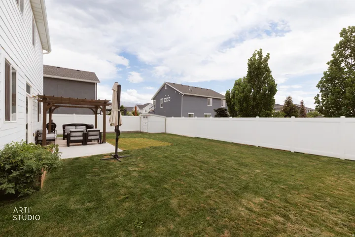 Expansive backyard featuring a manicured lawn, a concrete patio, a wooden pergola, a white vinyl fence, and a storage shed