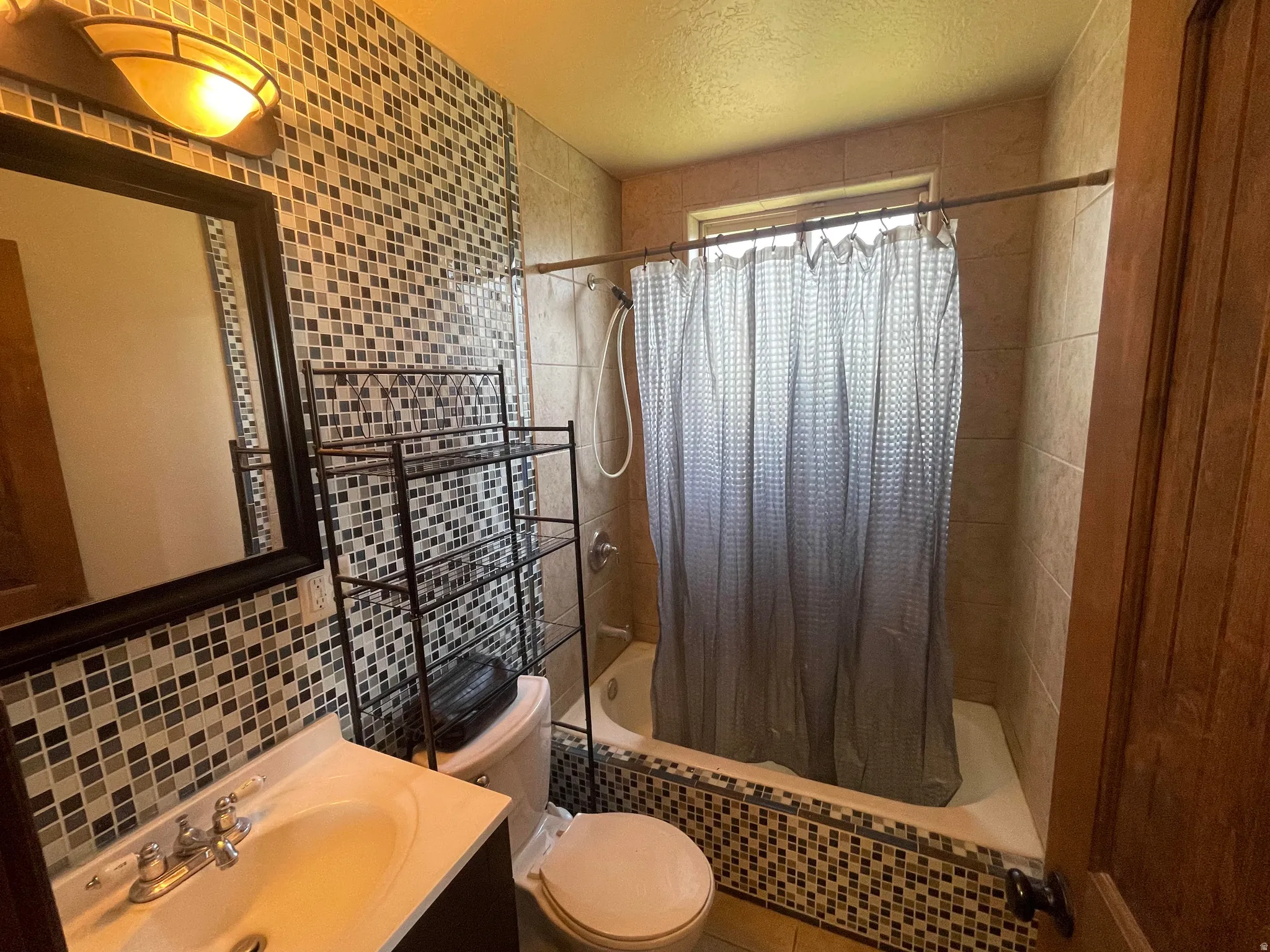 Spa-inspired bathroom featuring a walk-in shower with mosaic tile surround and glass enclosure, pedestal sink with chrome faucet, dual vanity with wood cabinetry, decorative tile backsplash, and a sheer curtain window treatment providing natural light