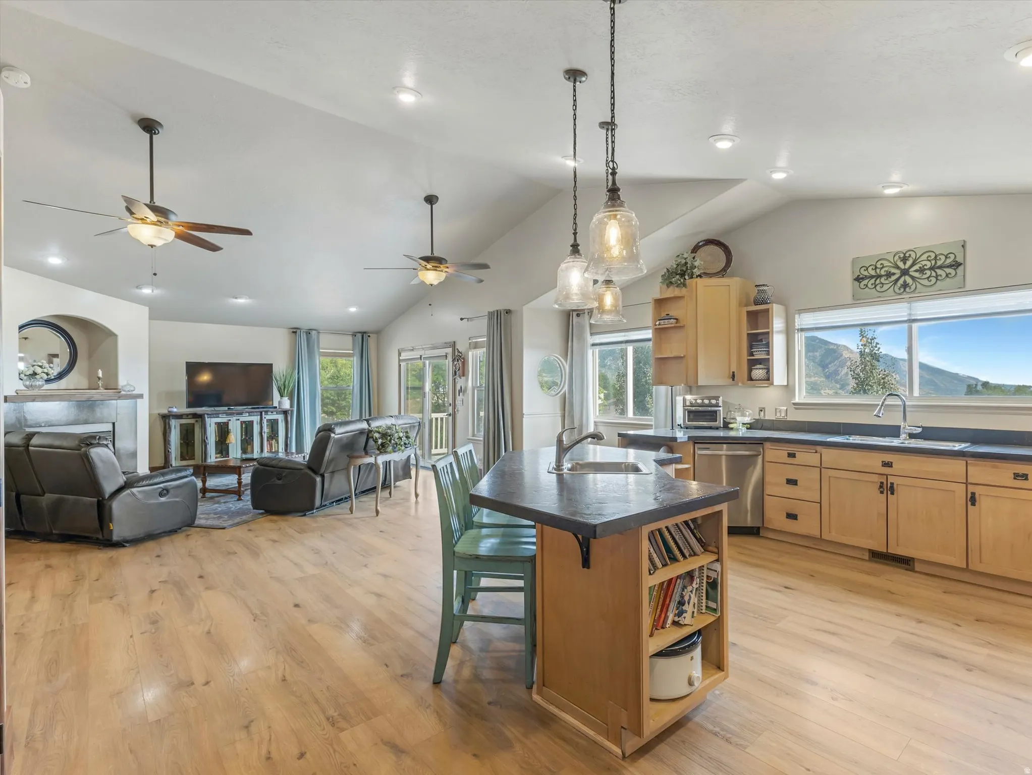 Open-concept living and kitchen area featuring wood-finish flooring throughout, a large center island with seating, custom cabinetry with ample storage, stainless steel appliances, pendant lighting, ceiling fans, recessed lighting, and expansive windows w