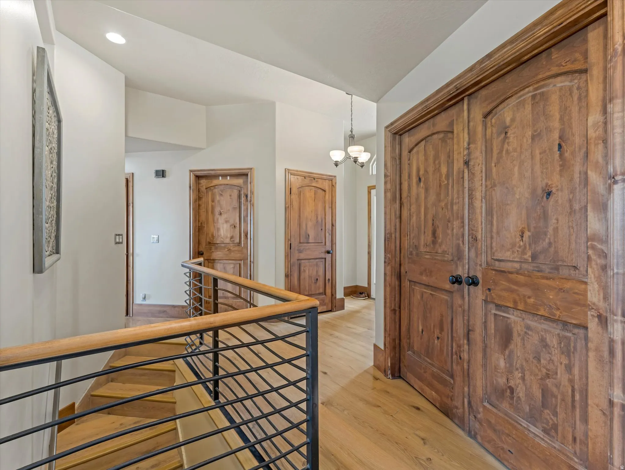 Hallway with double door to office/den, featuring warm wood-finish flooring, custom wood-panel doors with dark hardware, horizontal cable railing with wood top rail, recessed lighting, and white walls with clean architectural detailing