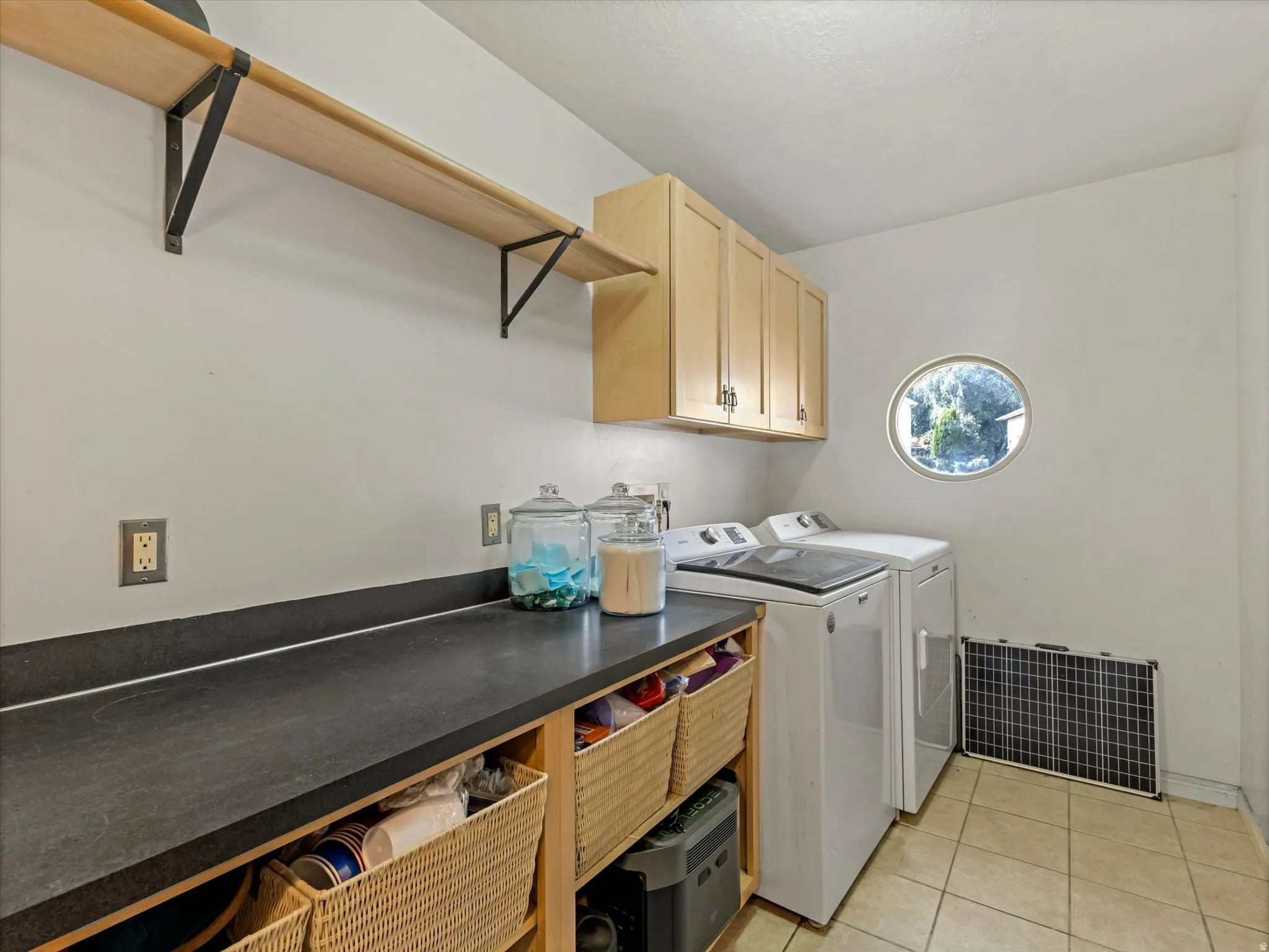Main Level Laundry,  featuring custom wood cabinetry with open storage cubbies, dark countertop workspace, stainless steel washer and dryer, tile flooring, and floating wood shelving with metal brackets