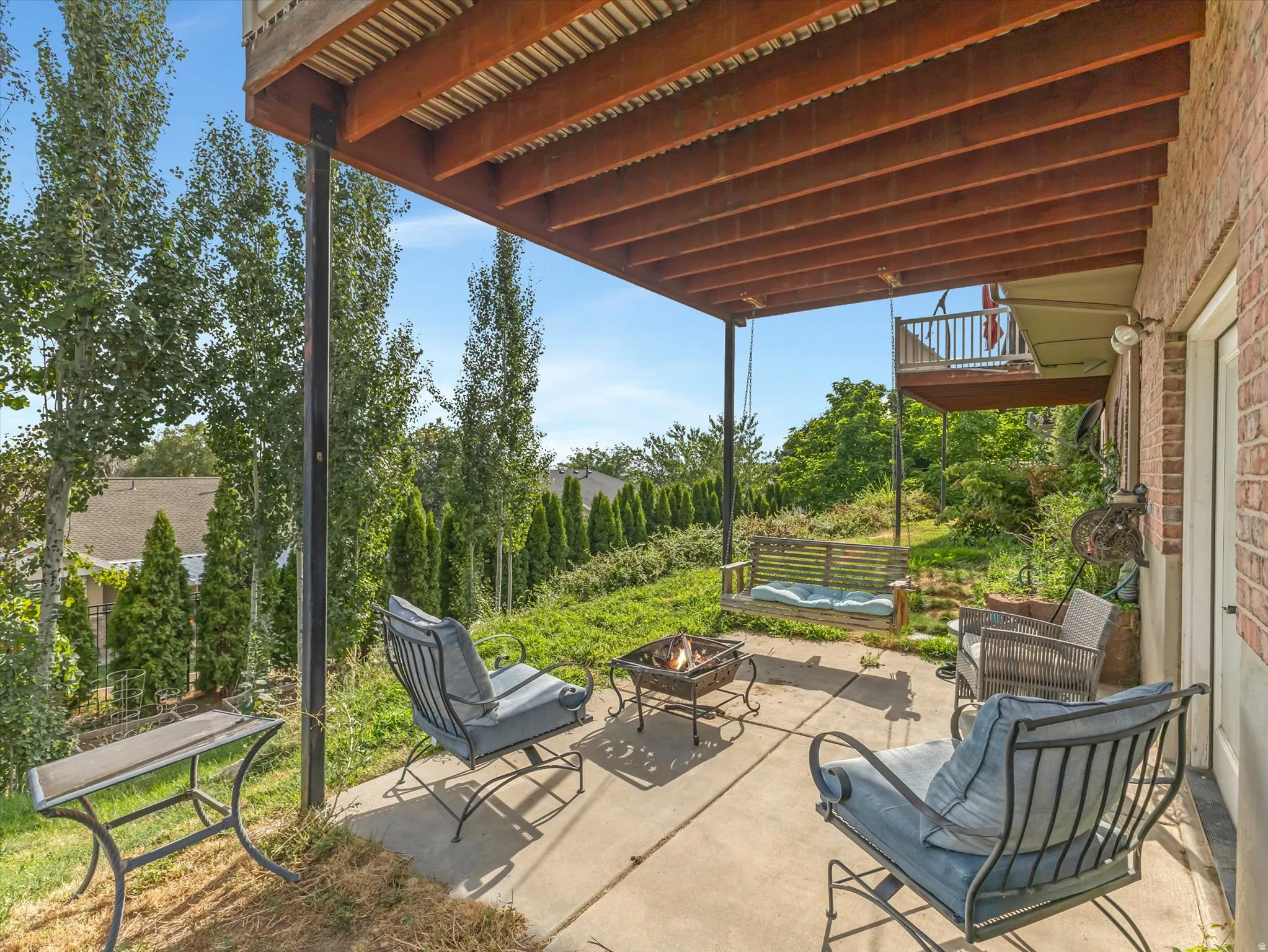 Covered patio with exposed wood beam ceiling and metal support posts overlooking manicured grounds featuring mature landscaping, ornamental evergreens, and a water feature