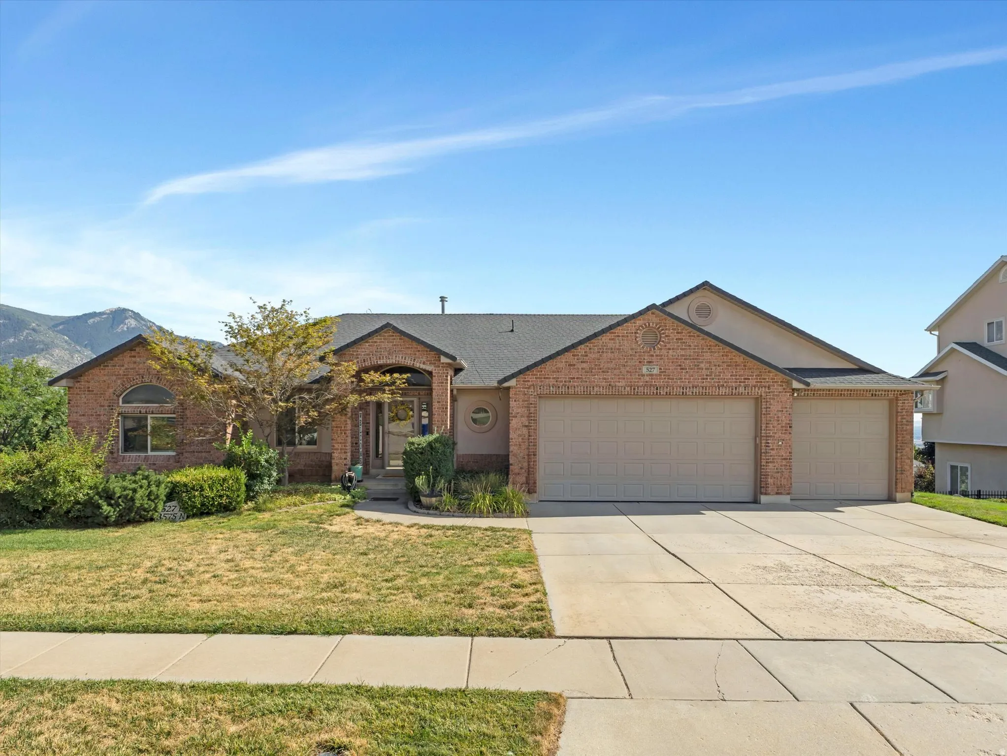 Brick Colonial home featuring a two-car garage, manicured front lawn with mature landscaping, concrete driveway, and mountain backdrop