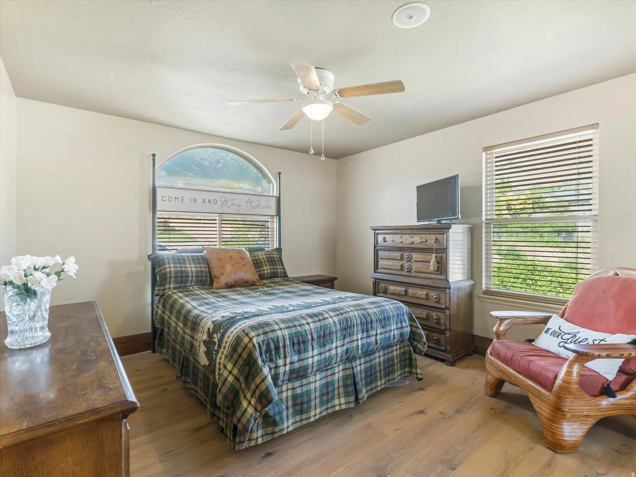 Bright secondary bedroom on main level featuring hardwood flooring, ceiling fan with integrated lighting, arched window with plantation shutters, and wood-finish dresser with integrated storage drawers