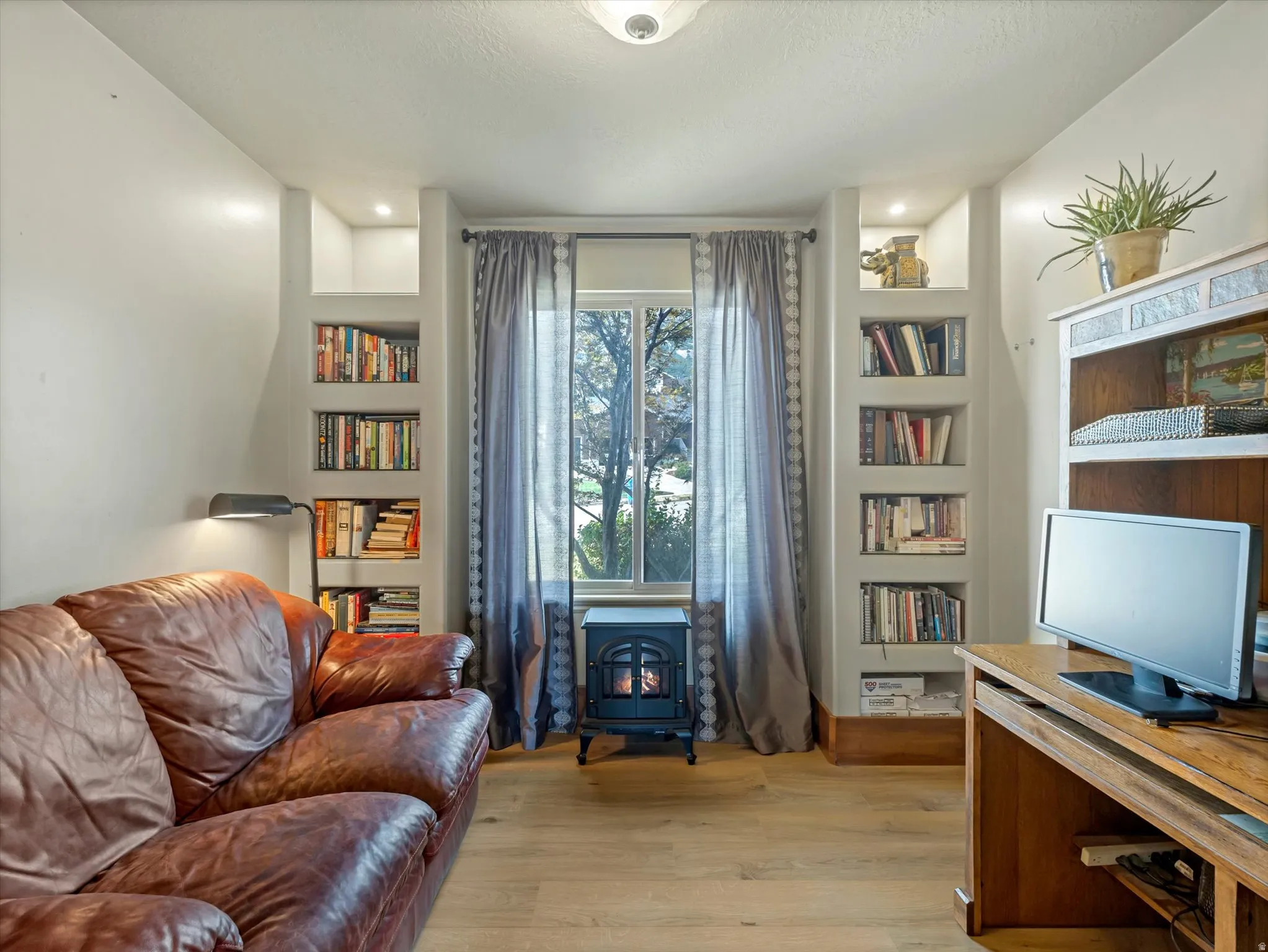Cozy den by front door, featuring built-in shelving throughout, wood-finish flooring, and a wood-burning stove centered beneath tall windows with gray drapery