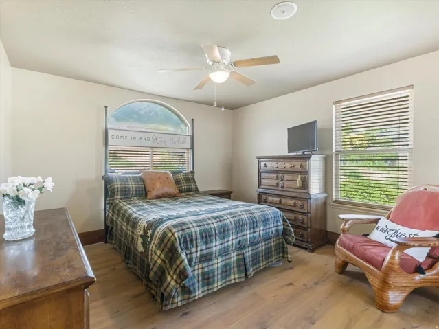 Bright secondary bedroom on main level featuring hardwood flooring, ceiling fan with integrated lighting, arched window with plantation shutters, and wood-finish dresser with integrated storage drawers