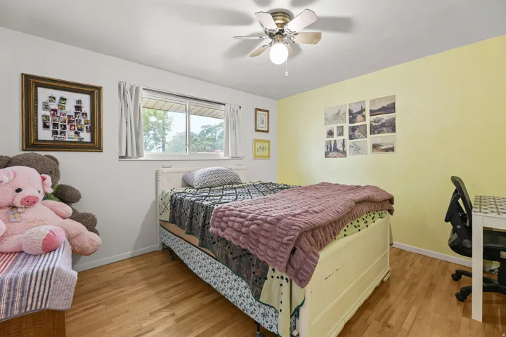 This bright bedroom features hardwood flooring, a ceiling fan, large window, and generous space for a bed and other furnishings.